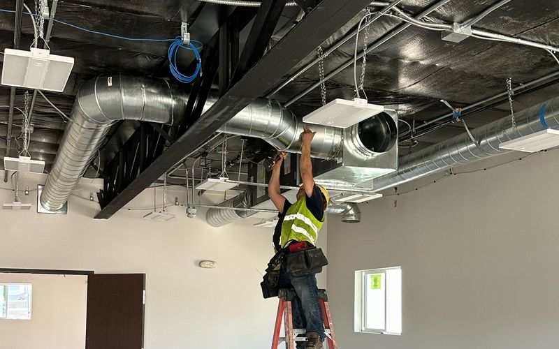 Installer on a ladder connecting spiral supply duct to a plenum in an open commercial interior
